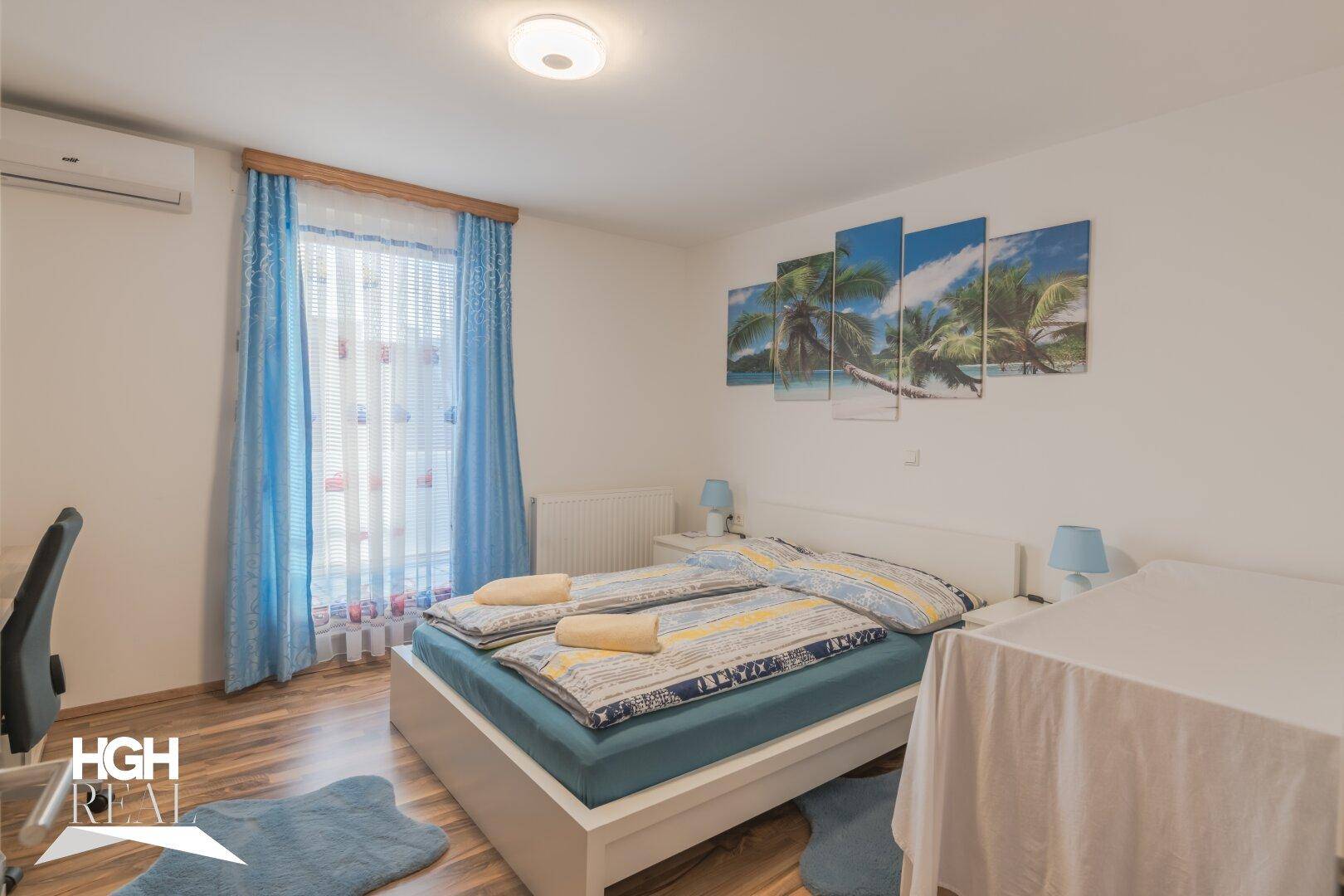 Helles Schlafzimmer mit Doppelbett, Holzboden und Klimaanlage. Blaue Vorhänge und Wandbilder schaffen eine entspannte Atmosphäre.
