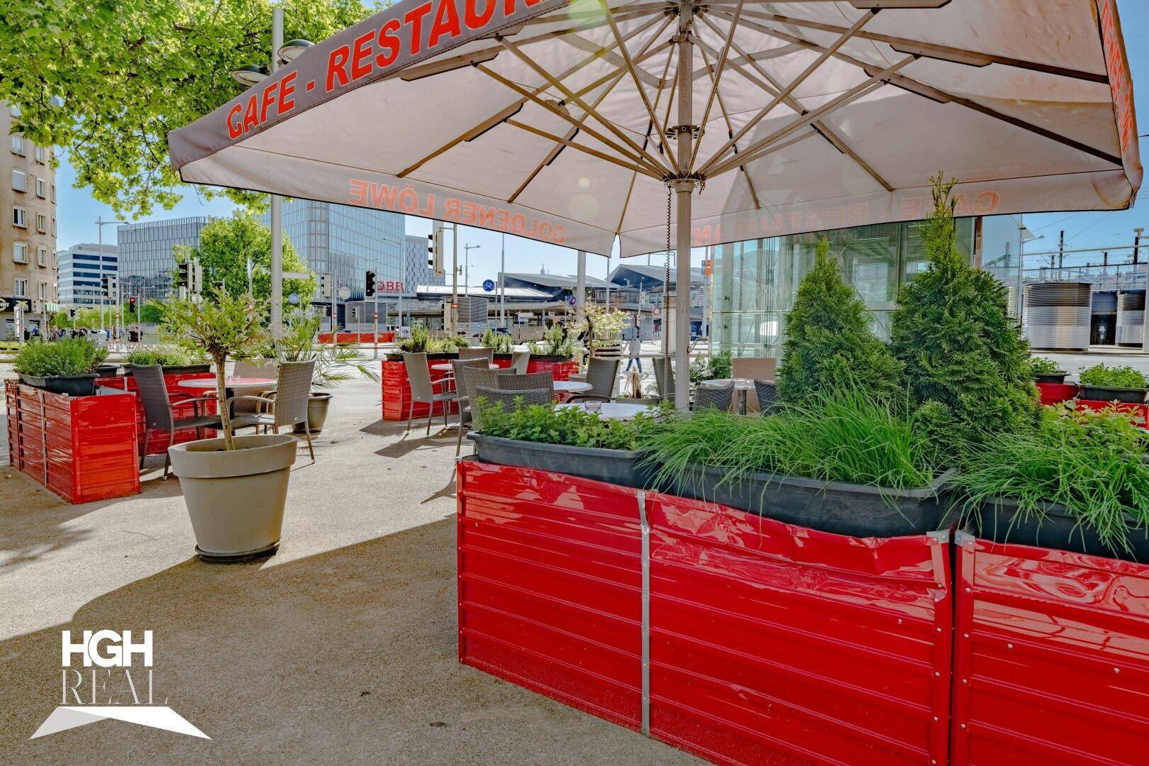 Sonnenverwöhnter Außenbereich des Cafés mit großen Schirmen, roten Pflanzkübeln und Sitzgelegenheiten auf dem Bürgersteig.