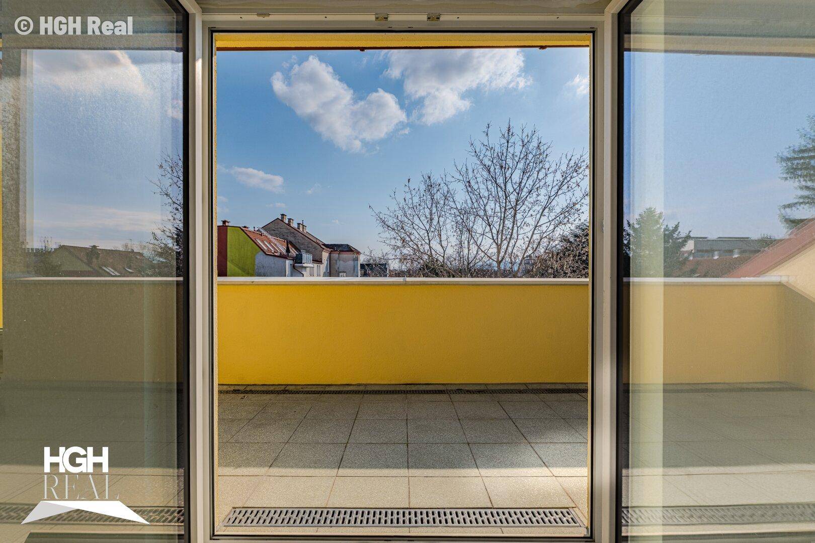 Blick von innen auf die geflieste Terrasse mit gelber Brüstung und Bäumen im Hintergrund.