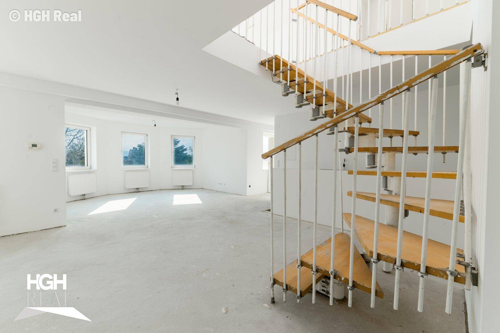 Lichtdurchfluteter, leerer Raum mit Betonboden und einer markanten Holztreppe, ideal für individuelle Einrichtung.