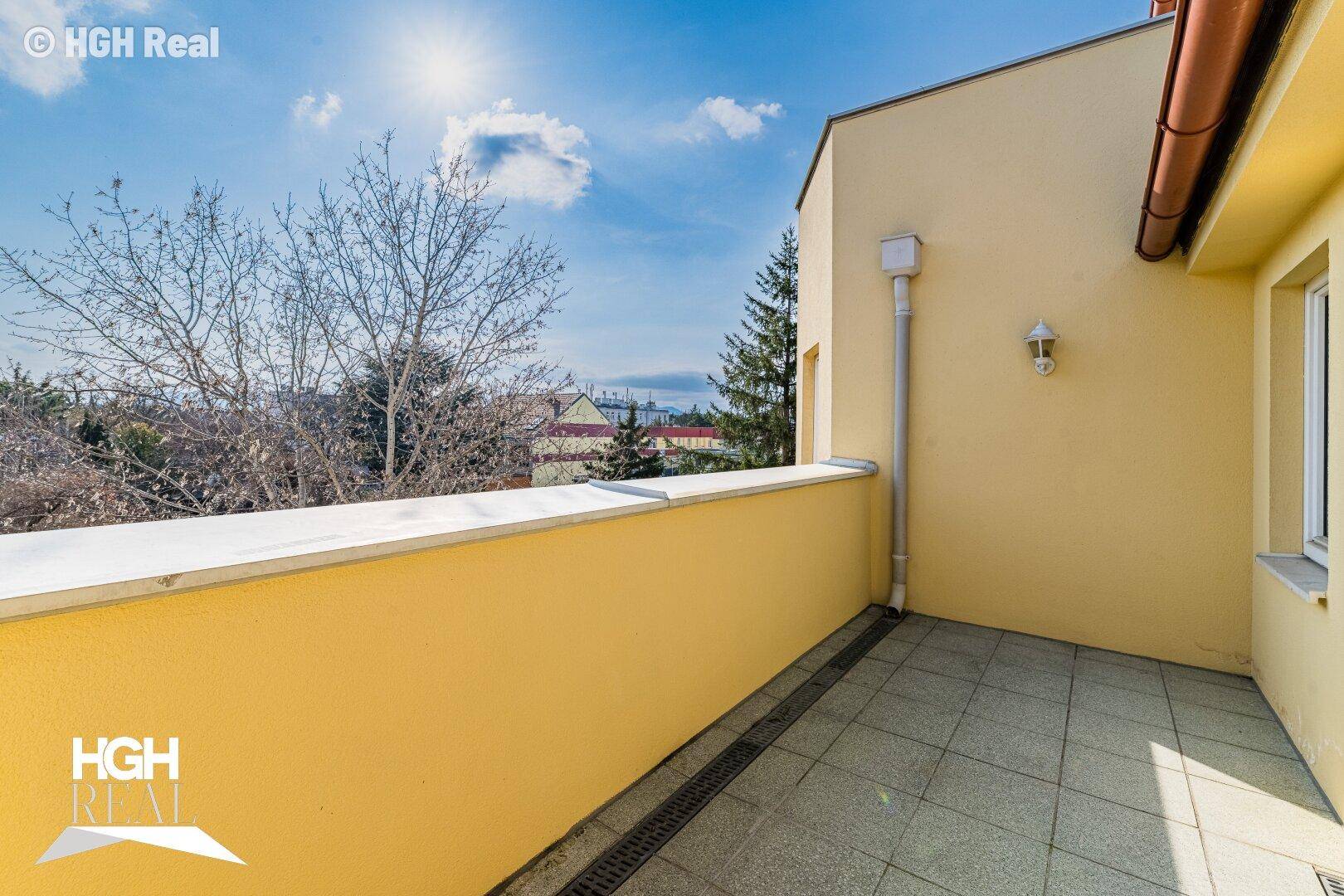 Sonniger Außenbereich mit gelber Brüstung, Fliesenboden und Blick auf Bäume und entfernte Gebäude.