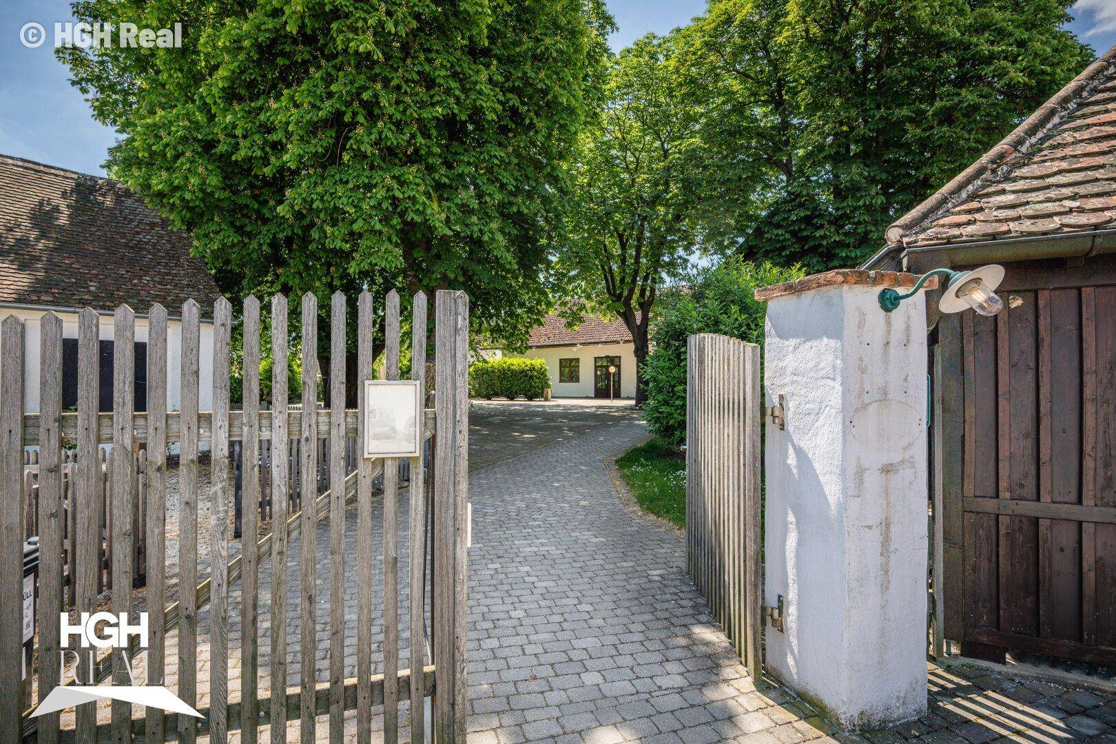 Offenes Holztor führt zu einem gepflasterten Weg, der zum Anwesen mit viel Grün führt.