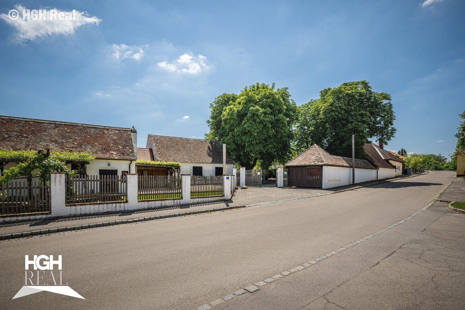 Ansicht des Anwesens von der Straße aus, mit Gebäuden, Zaun und Bäumen unter blauem Himmel.
