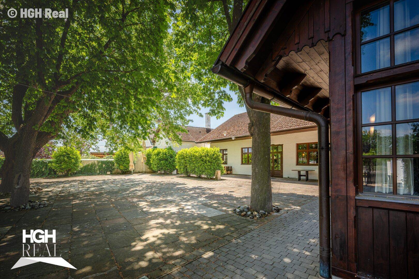 Großer gepflasterter Innenhof mit alten Bäumen und einem Gebäude im Hintergrund.