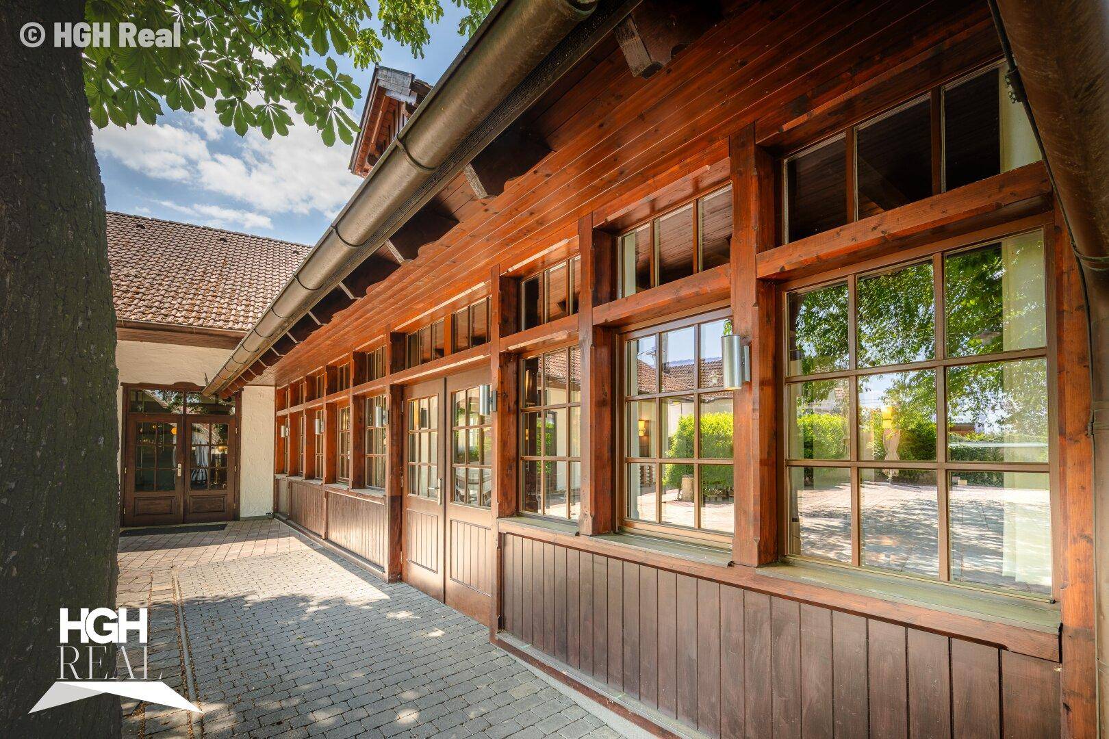 Gebäude mit Holzfassade und vielen Fenstern, daneben ein gepflasterter Weg unter Bäumen.