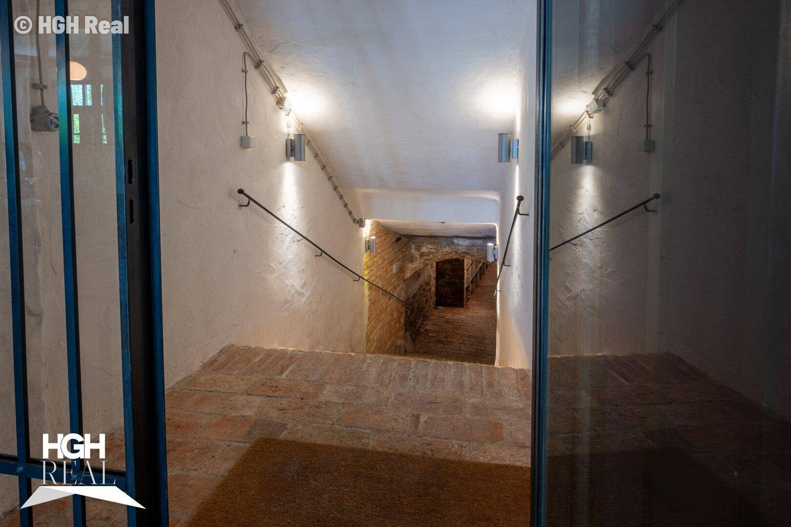 Treppenabgang zum Keller mit Ziegelboden und Handlauf, beleuchtet von Wandlampen.