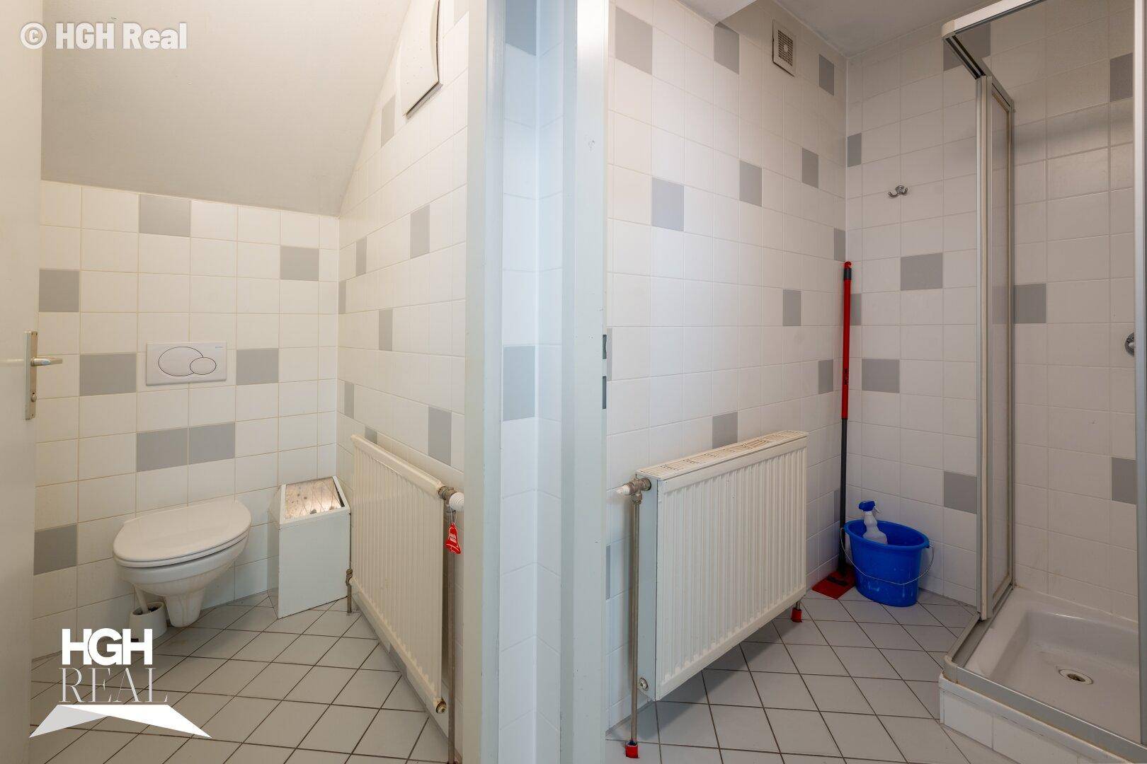 Einfaches Badezimmer mit Dusche, Toilette und Heizkörpern, gefliest in Weiß und Grau.