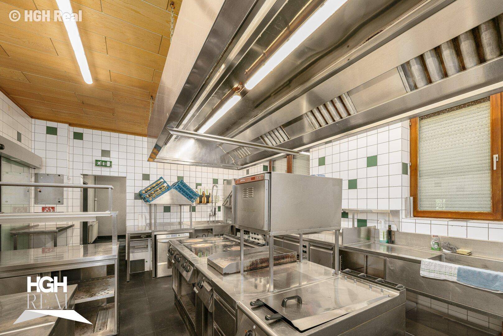 Professionelle Küche mit umfangreicher Edelstahlausstattung, Dunstabzugshaube und gefliesten Wänden.