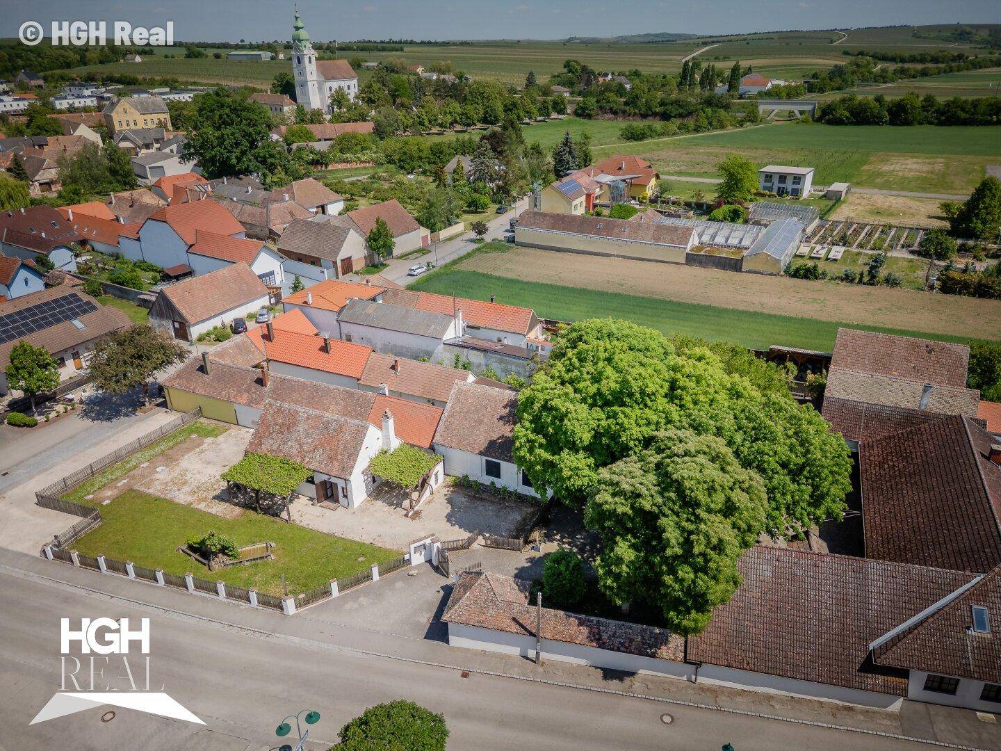 Weitwinkel-Luftaufnahme des Anwesens, eingebettet in ein Dorf mit Feldern und Kirche im Hintergrund.