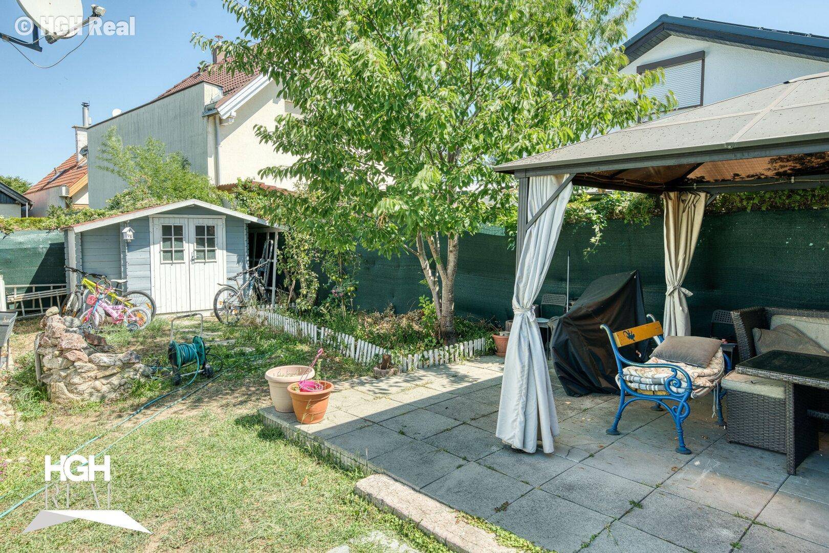 Gepflegter Gartenbereich mit Gartenhaus, gepflasterter Terrasse und einem schattenspendenden Baum.