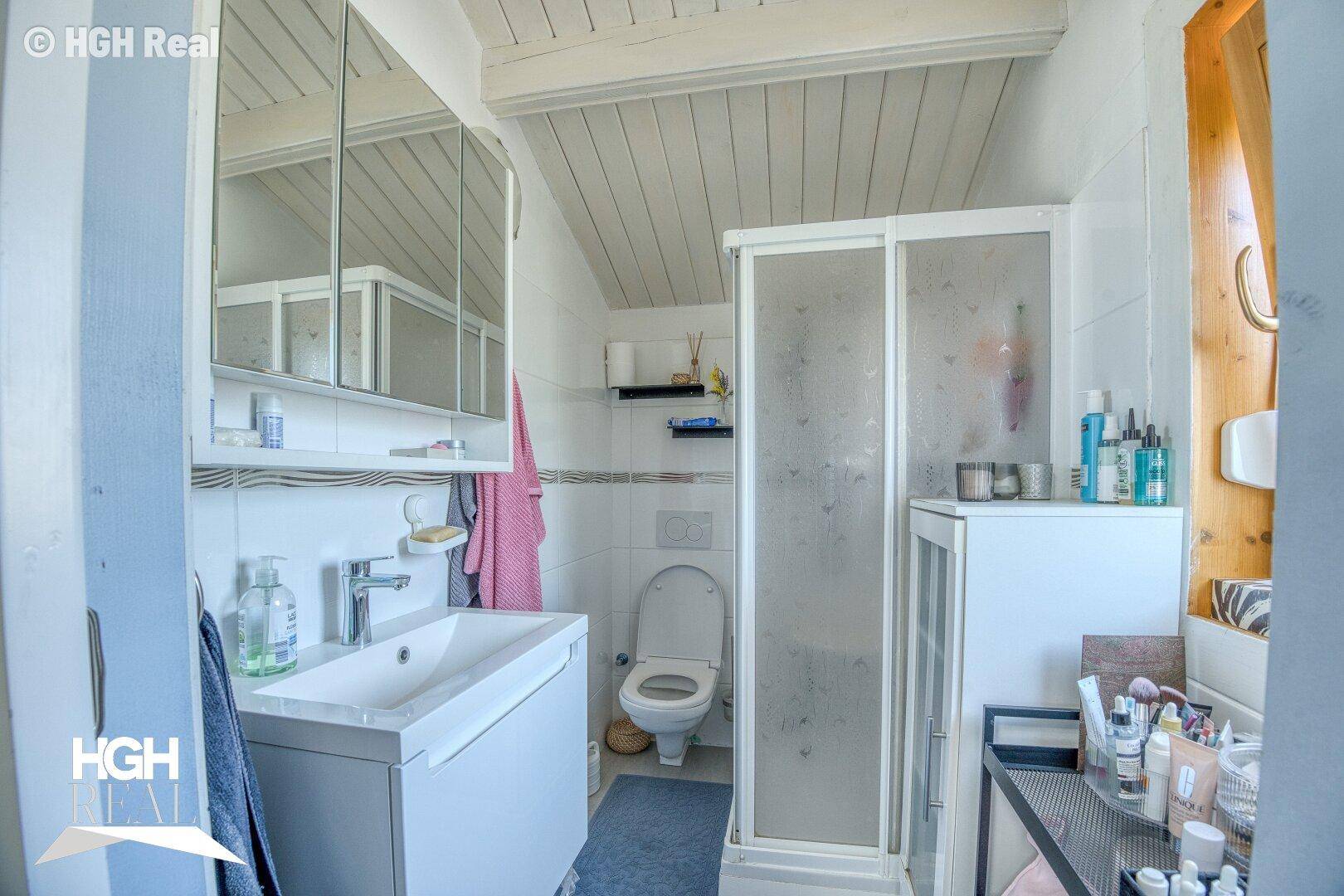 Helles Badezimmer mit weißen Fliesen, Duschkabine, Einzelwaschbecken und Dachschräge.