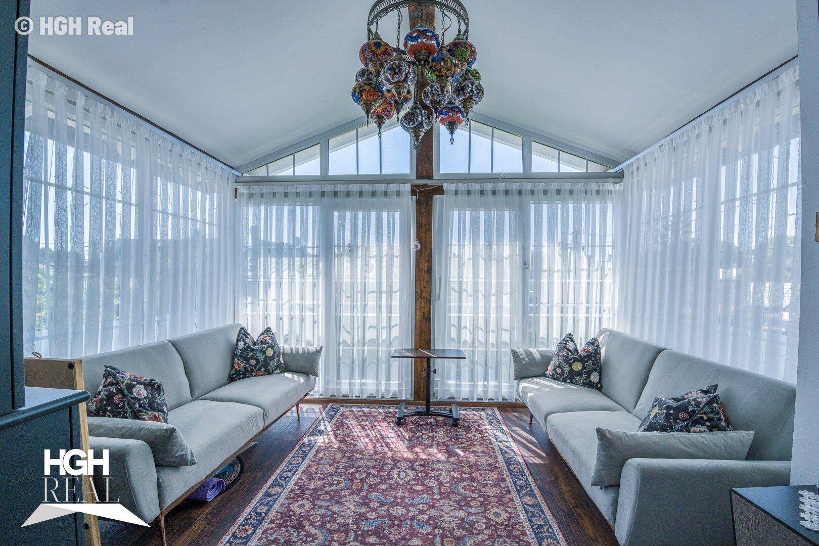 Gemütlicher Wintergarten mit zwei Sofas, Teppich und einem farbenfrohen Kronleuchter, umgeben von Fenstern.