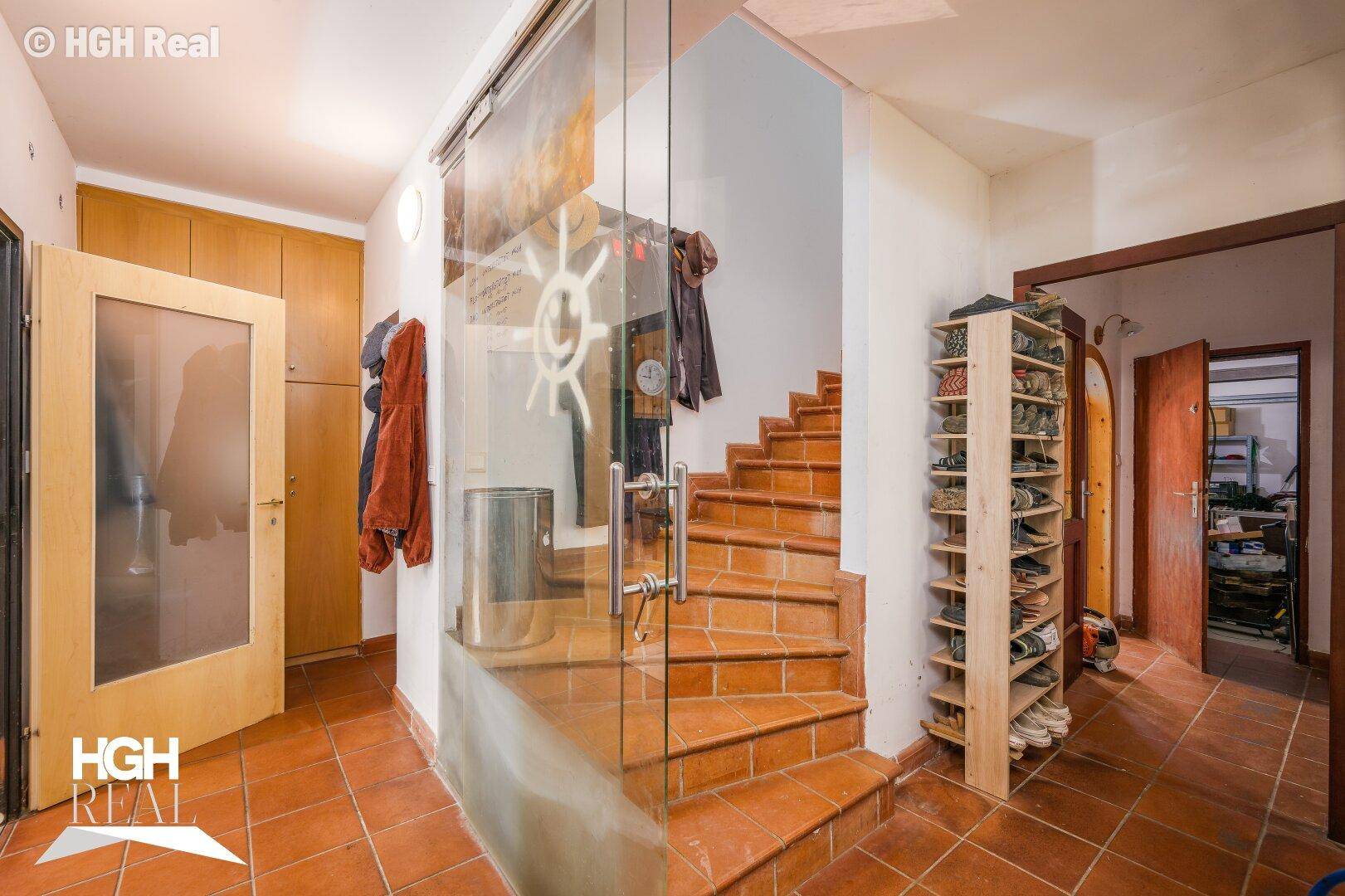 Eingangsbereich mit Terrakottafliesen, Holztreppe und einer Glastür.
