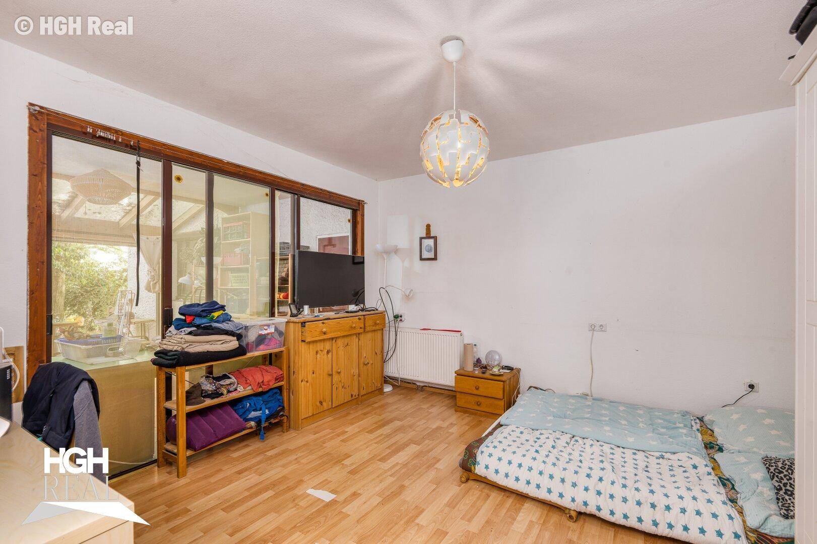 Schlafzimmer mit Laminatboden, Fenster zum Wintergarten und einfacher Einrichtung.