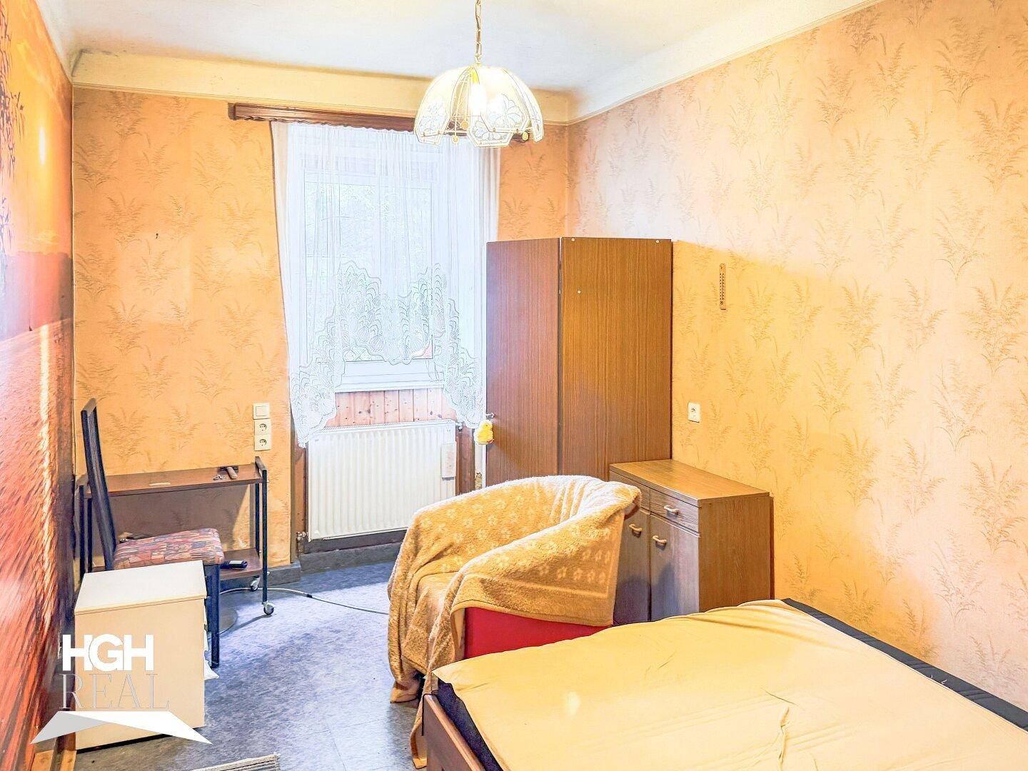 Schlafzimmer mit gemusterter Tapete, einfachem Mobiliar und einem Fenster mit Blick ins Grüne.