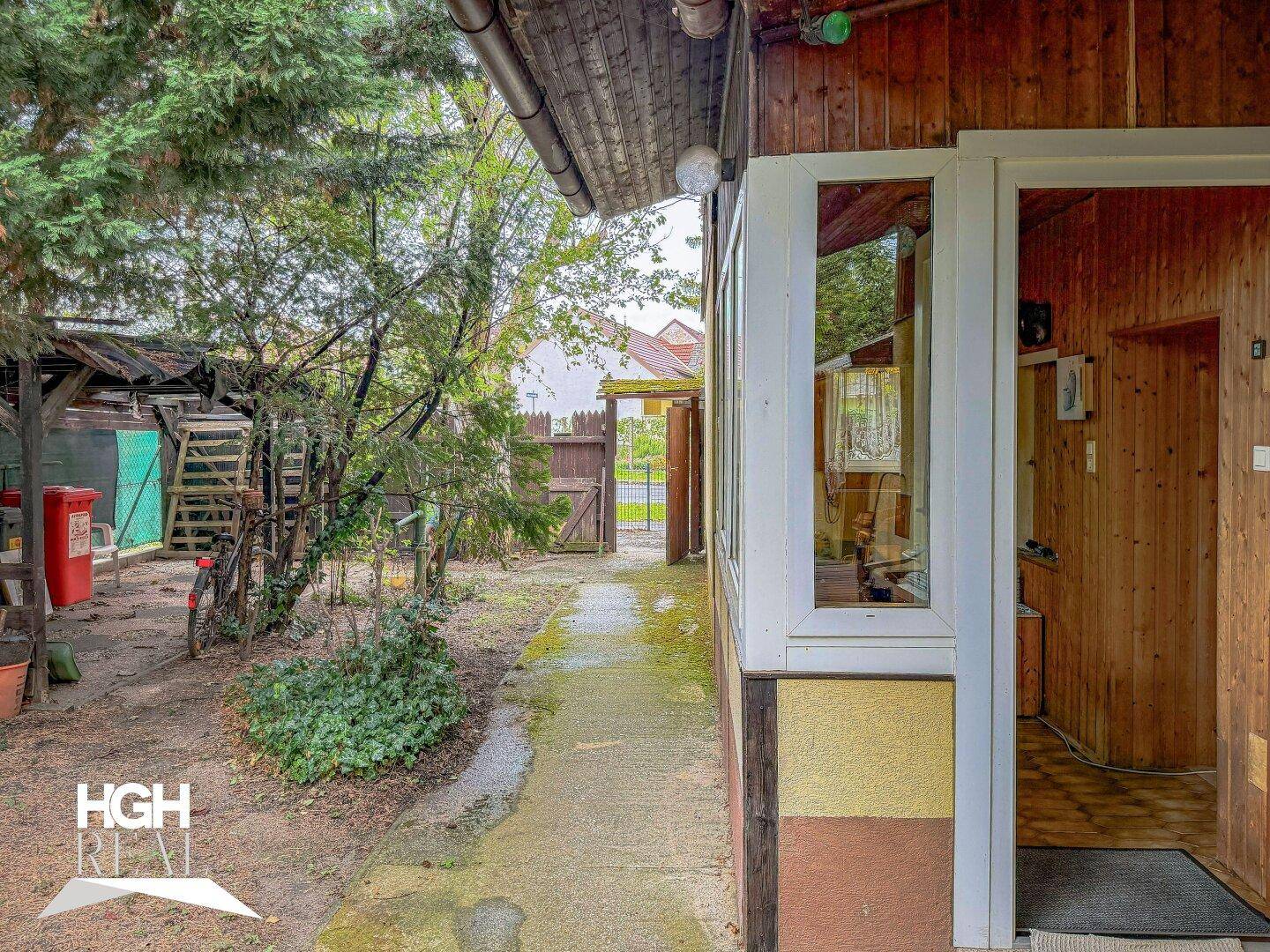 Schmaler Weg entlang des Hauses mit Holzwänden und Blick auf ein Gartentor im Hintergrund.