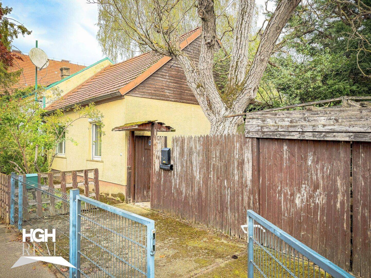 Älteres Einfamilienhaus mit gelber Fassade, Holzzaun und Eingangsbereich, umgeben von Bäumen.