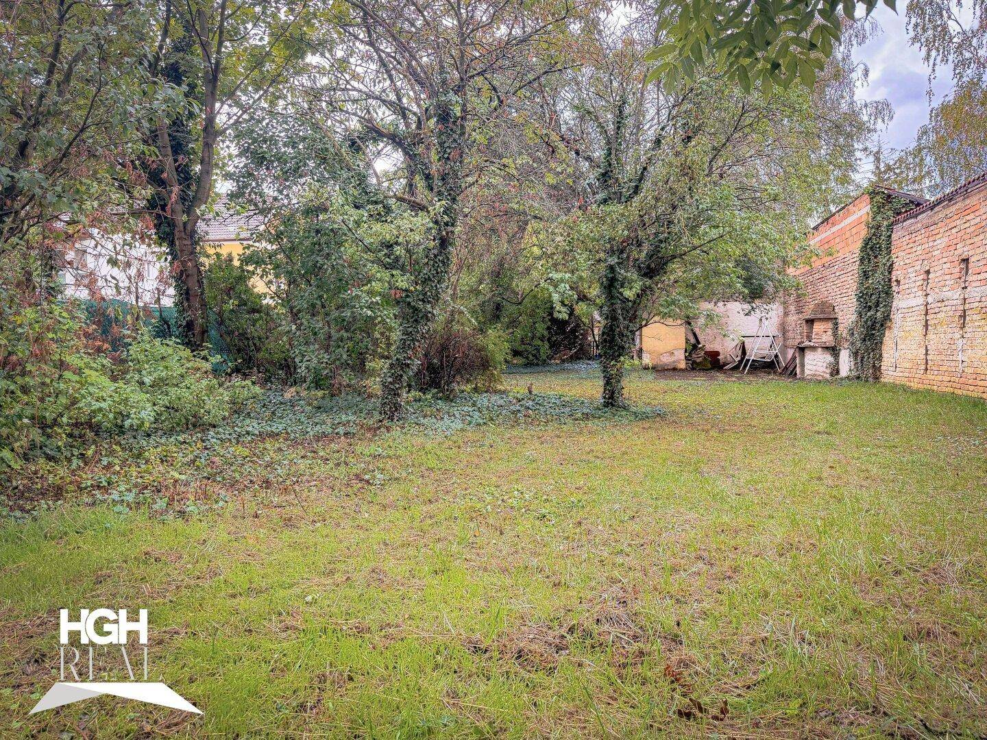 Weitläufiger Gartenbereich mit Rasenfläche, alten Bäumen und einer Backsteinmauer im Hintergrund.