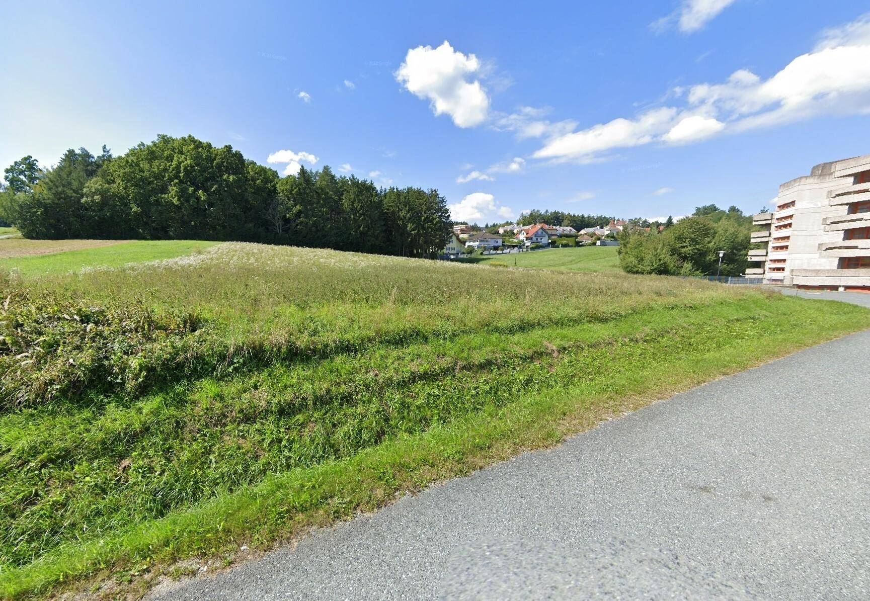 Offenes Feld mit grüner Vegetation, umgeben von Bäumen und Häusern in der Ferne.