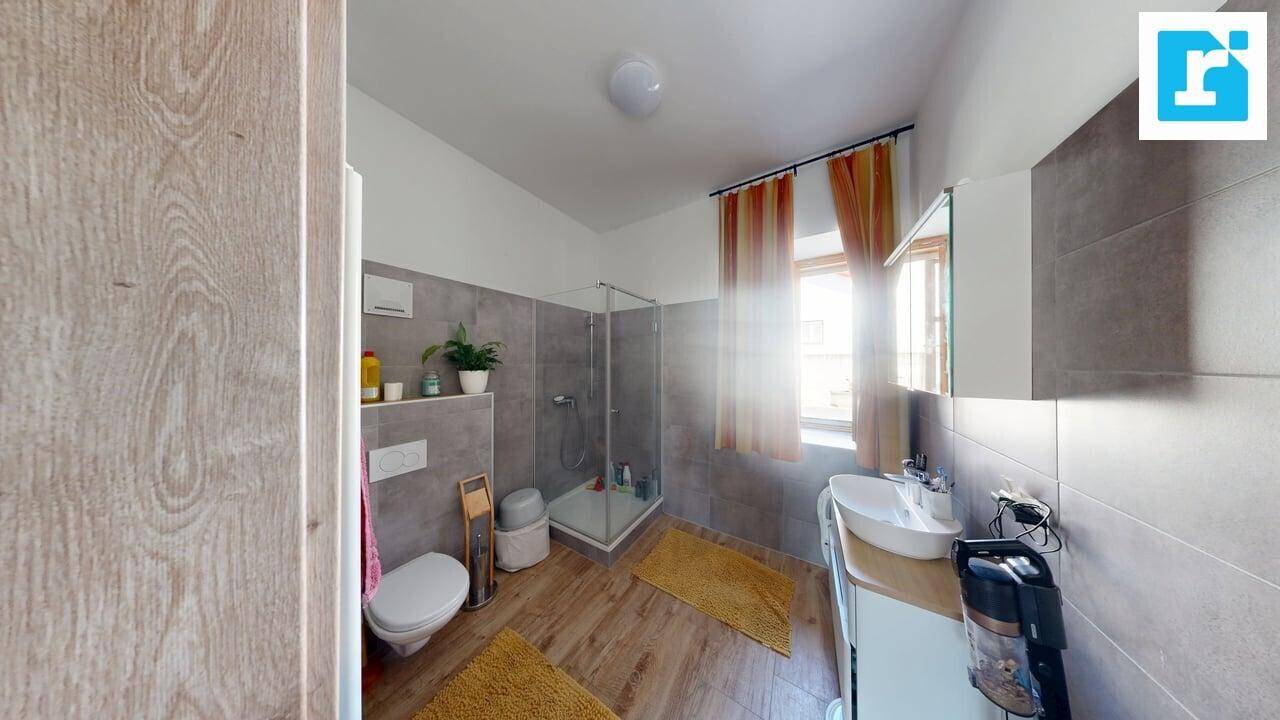 Badezimmer mit grauen Fliesen, Holzboden, Duschkabine und WC, hell durch das Fenster mit orangefarbenen Vorhängen.