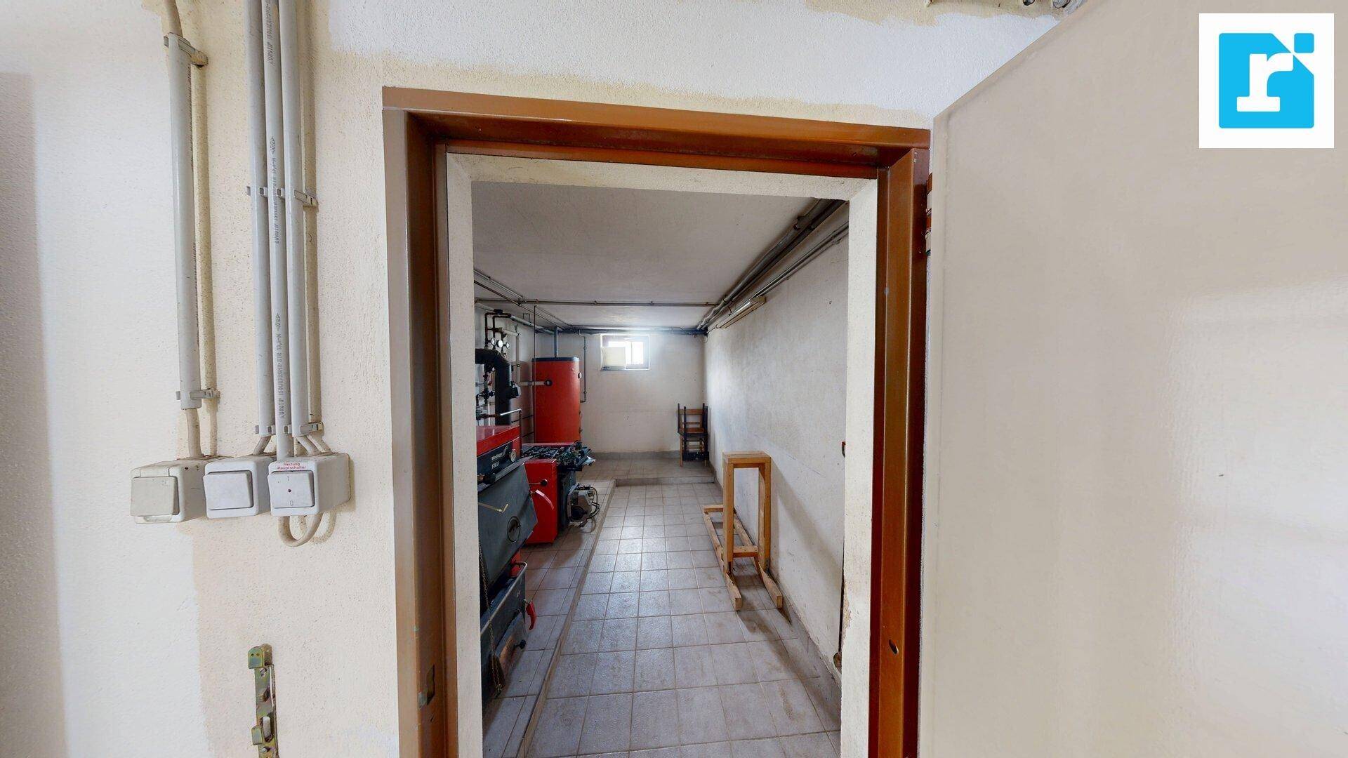 Blick in einen Heizungsraum im Keller mit rotem Boiler, gefliestem Boden und sichtbaren Installationen.