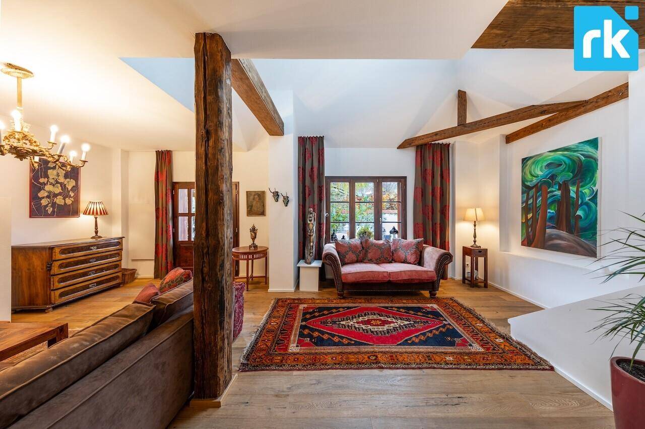 Geräumiges Wohnzimmer mit hohen Decken, sichtbaren Holzbalken und großen Fenstern, sehr hell.