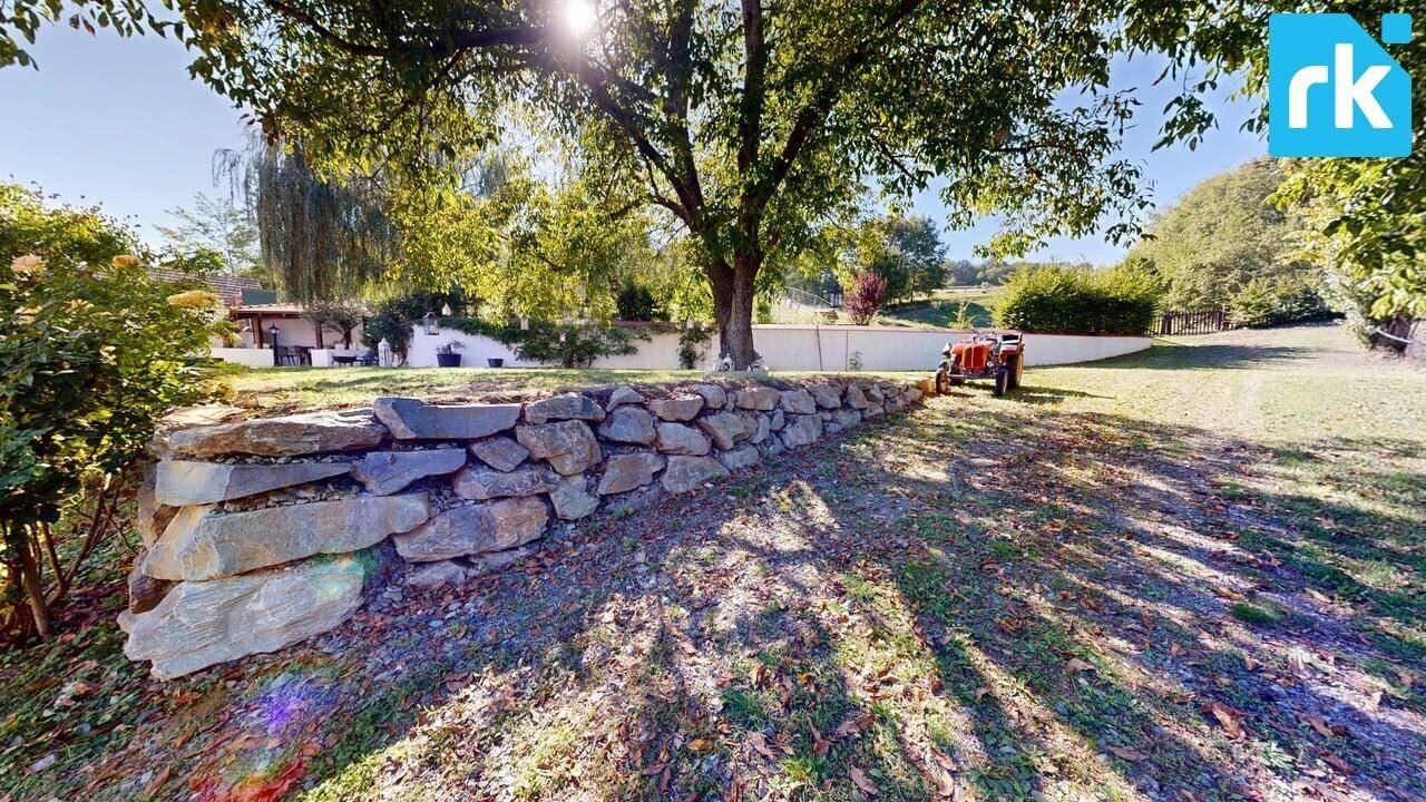 Sonniger Gartenbereich mit Natursteinmauer, Rasenfläche und Bäumen im Hintergrund.