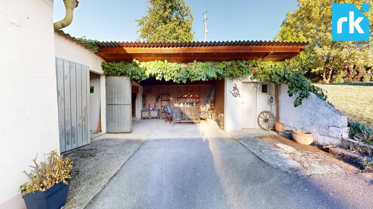 Überdachter Stellplatz oder Carport mit Holzbalken und Rankpflanzen, daneben ein Lagerbereich.