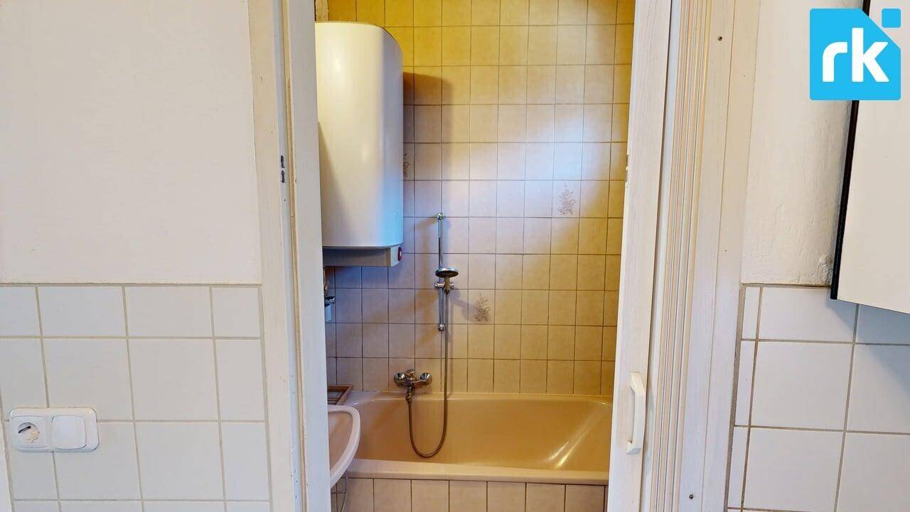 Kleines Badezimmer mit Badewanne, beigefarbenen Fliesen und einem Warmwasserboiler.