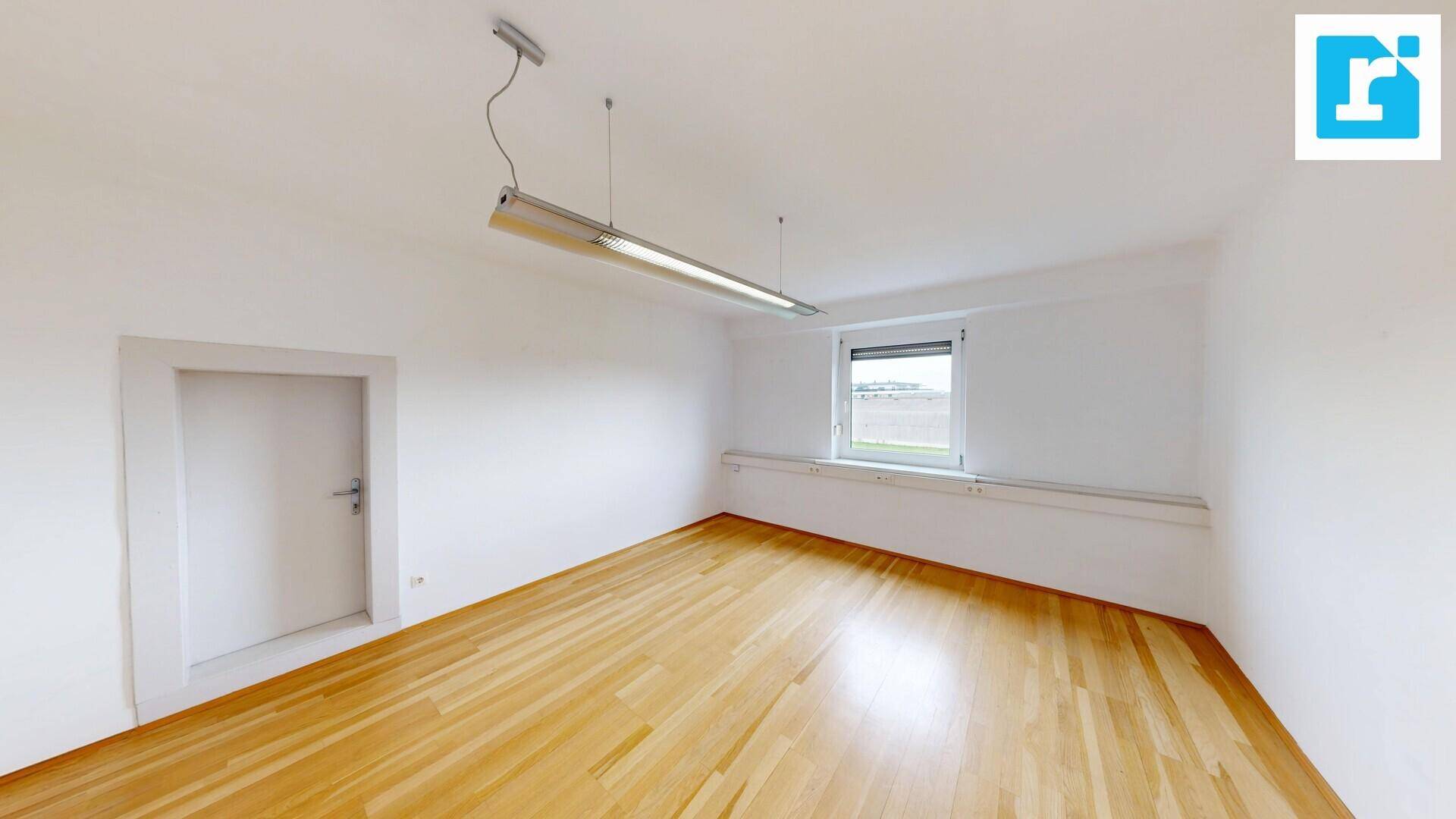 Leeres Büro mit Holzboden, weißen Wänden und einem Fenster mit Rollladen, bereit zur individuellen Gestaltung.