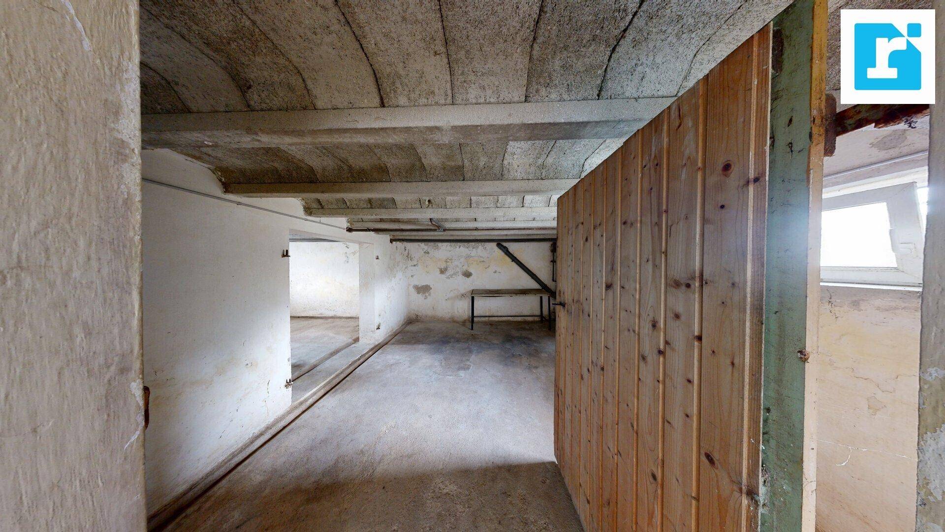 Langer Kellerraum mit Betonboden, unverputzten Wänden und einer Holztür, die zu weiteren Räumen führt.