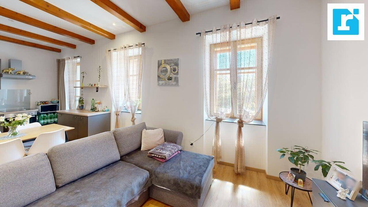Gemütlicher Wohnbereich mit grauem Ecksofa, Holzboden und sichtbaren Holzbalken an der Decke.