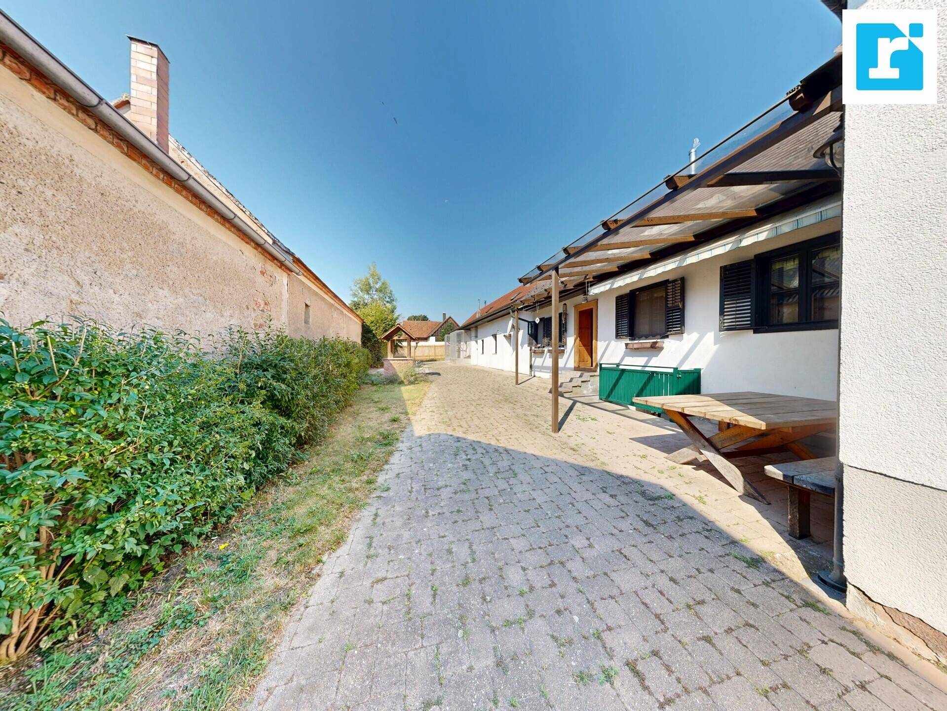 Langer gepflasterter Weg entlang der Hausfassade mit grünen Büschen und Sitzgelegenheiten.