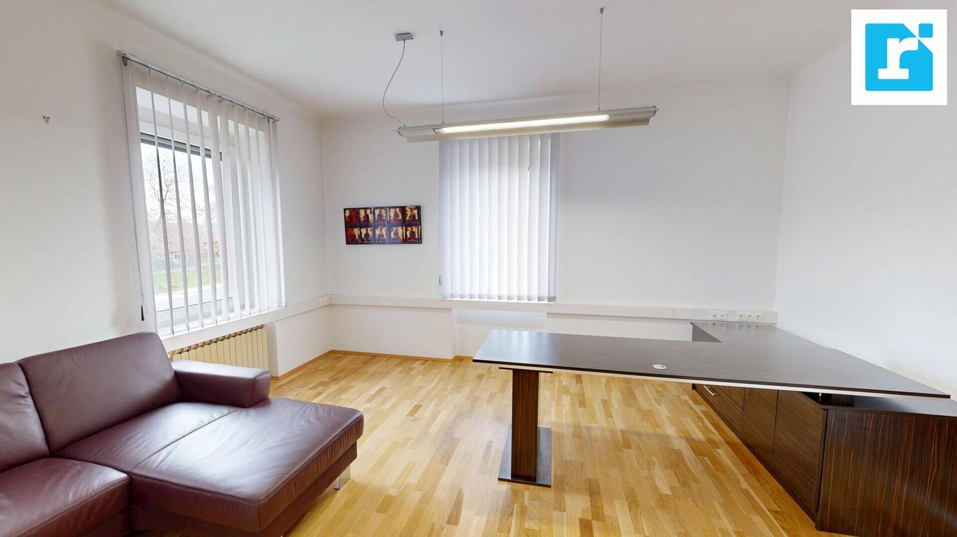 Geräumiges Büro mit Holzboden, einem großen Schreibtisch und einer bequemen Ledercouch, gut beleuchtet durch Fenster.