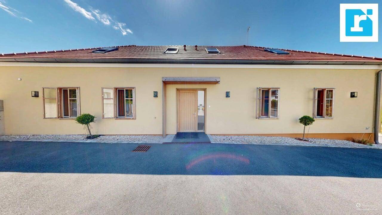 Ansicht des Hauses mit heller Fassade, braunem Ziegeldach und mehreren Fenstern an einem sonnigen Tag.