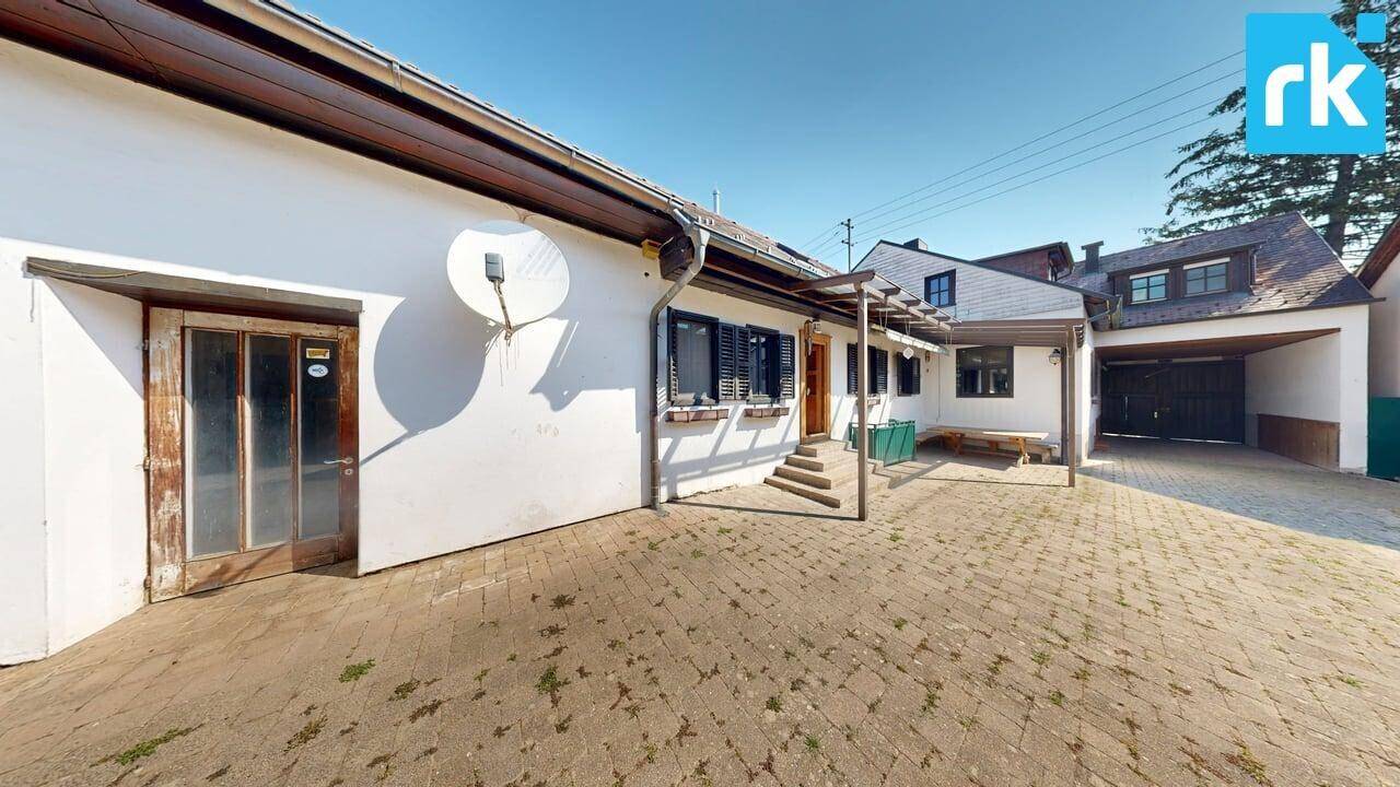 Hausfassade mit gepflastertem Vorplatz und einem überdachten Carport im Hintergrund.