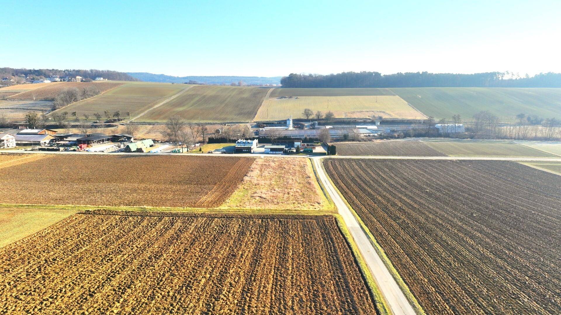 Luftaufnahme von Feldern und einer Straße mit Gebäuden und sanften Hügeln in der Ferne.