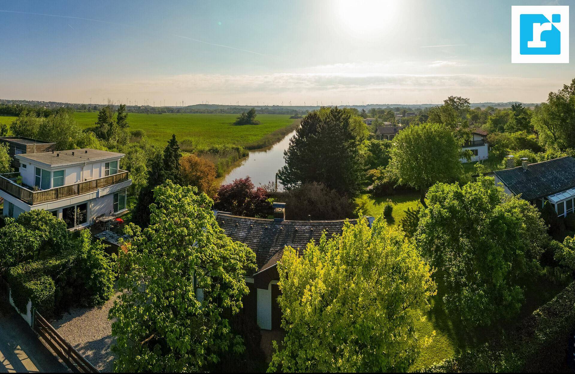 Luftaufnahme einer Wohnsiedlung am Wasser mit weiten Feldern und viel Grün unter blauem Himmel.