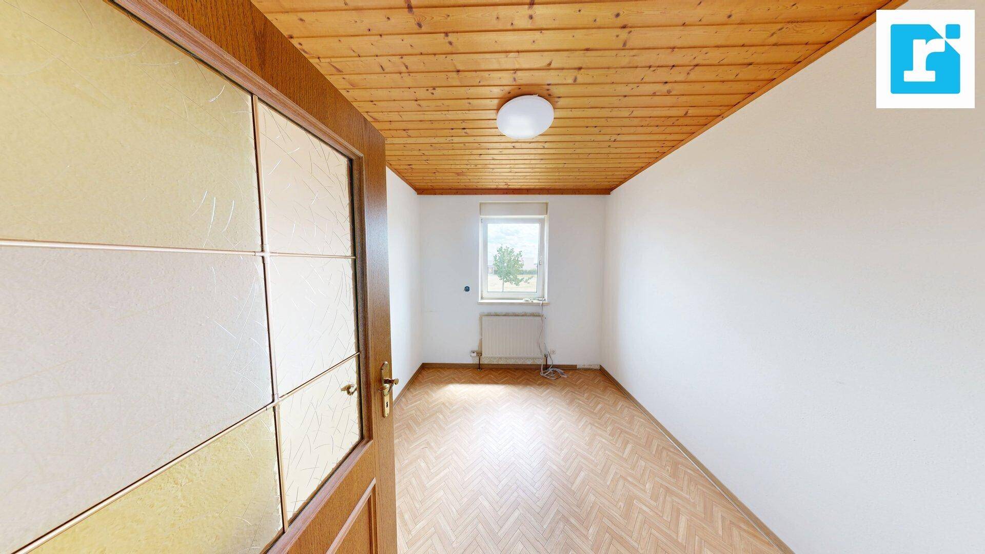 Leeres Zimmer mit Holzdecke und Parkettboden, hell durch ein Fenster mit Ausblick.