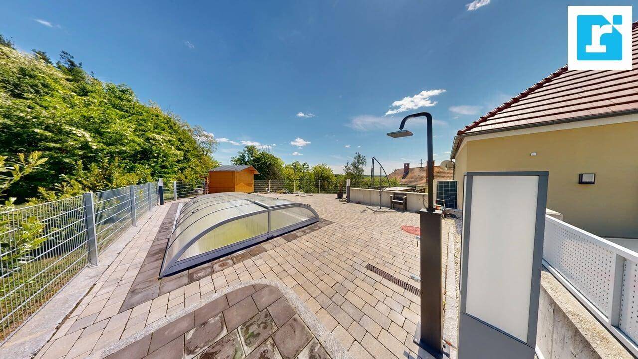 Gepflasterter Außenbereich mit überdachtem Pool, Außendusche und Gartenhaus, umgeben von Grün.
