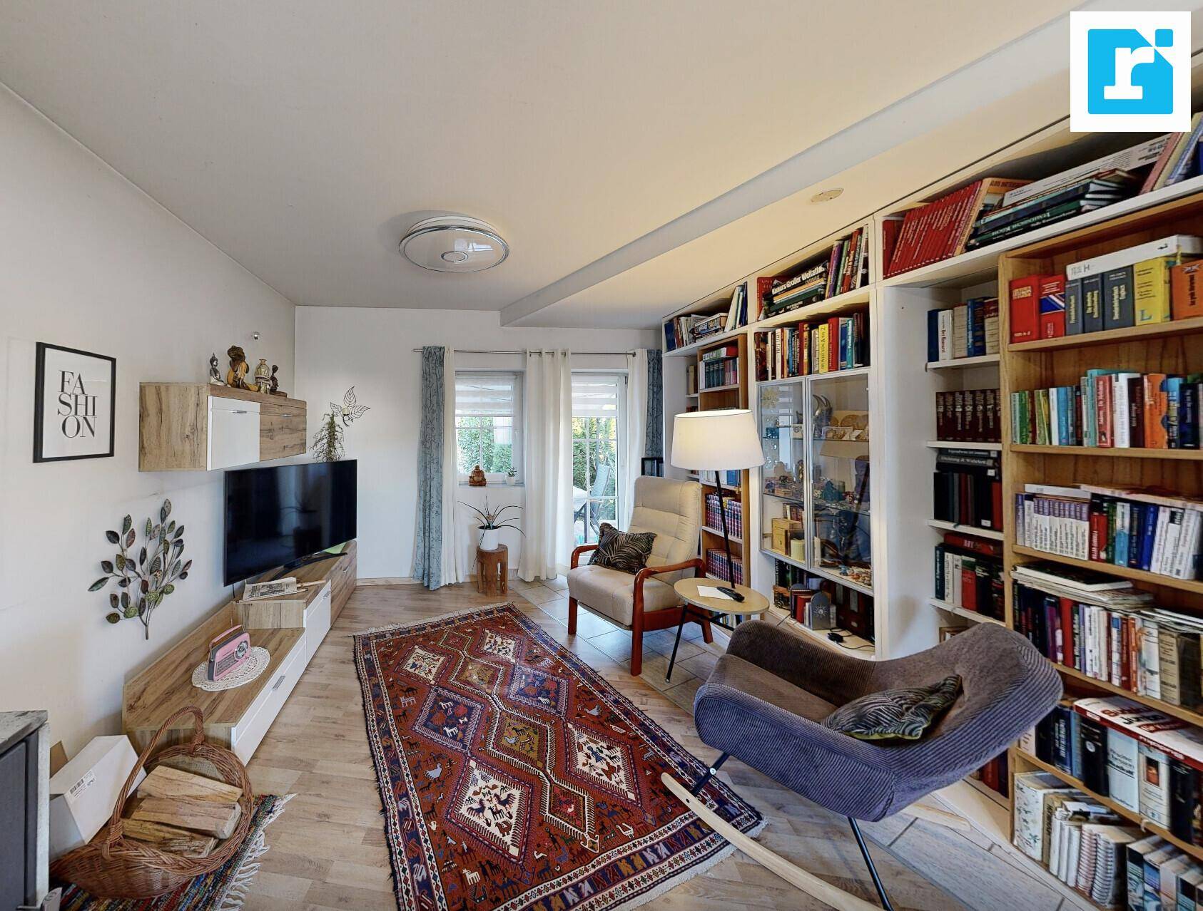 Helles Wohnzimmer mit Fernseher, Bücherregal und Teppich auf Laminatboden.
