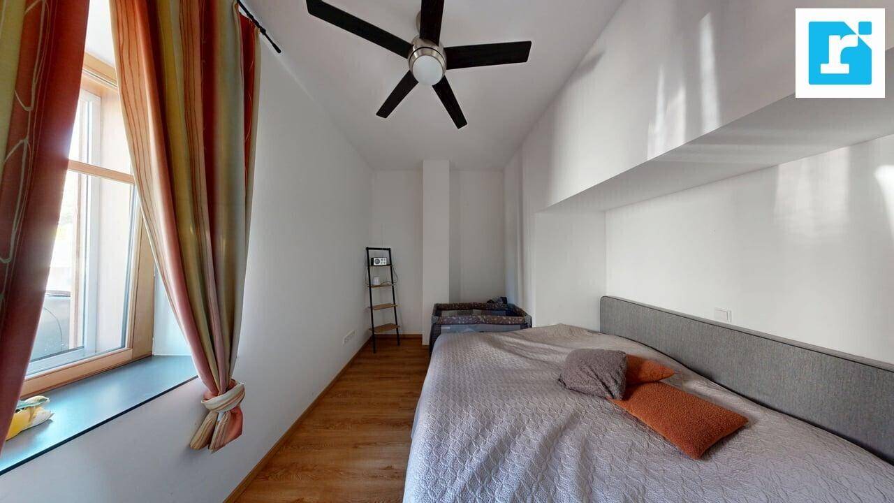 Schlafzimmer mit Holzboden, weiß gestrichenen Wänden, Fenster mit Vorhängen und Deckenventilator.