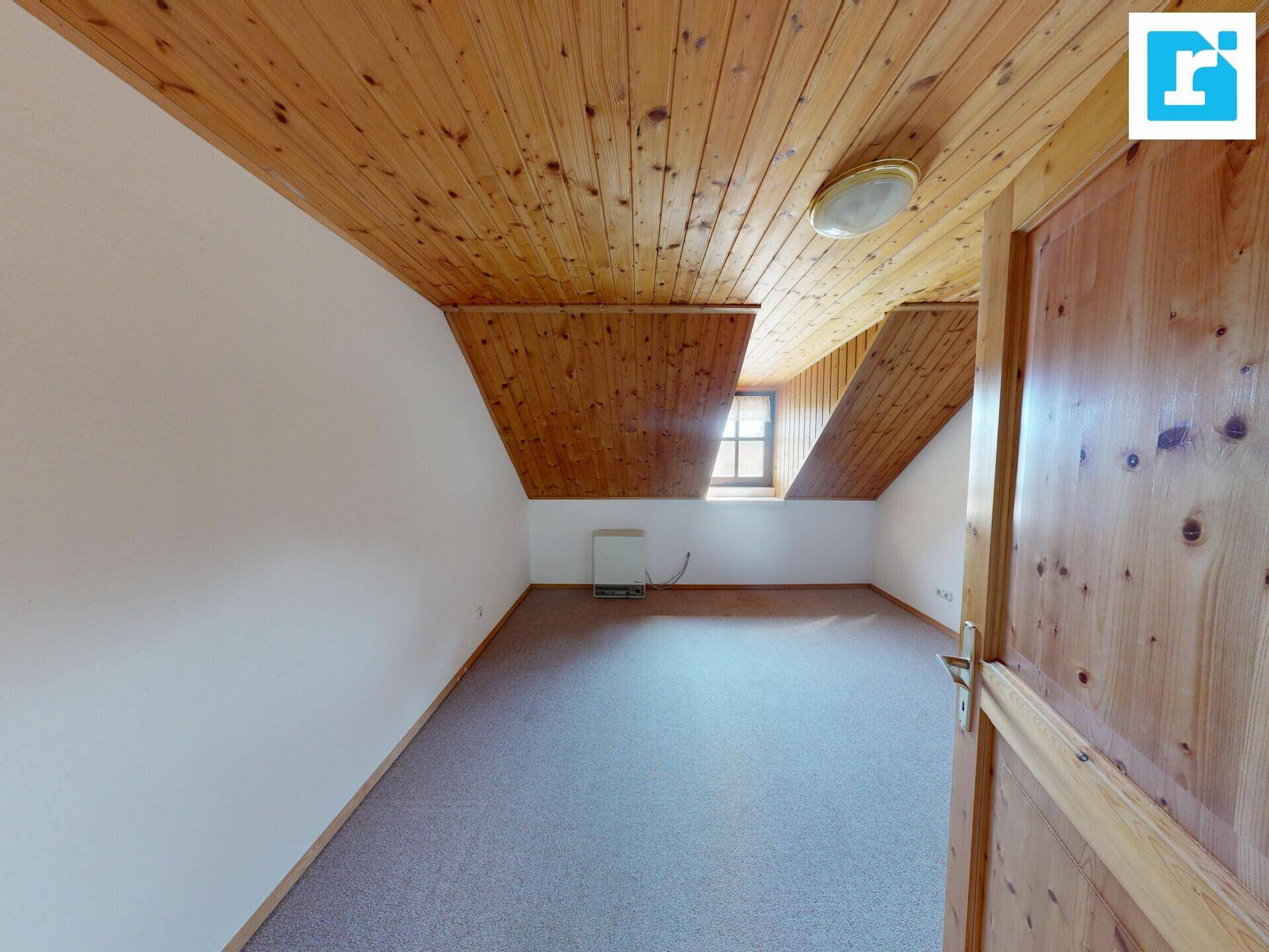 Leeres Dachzimmer mit Holzdecke, Teppichboden und einem kleinen Fenster.