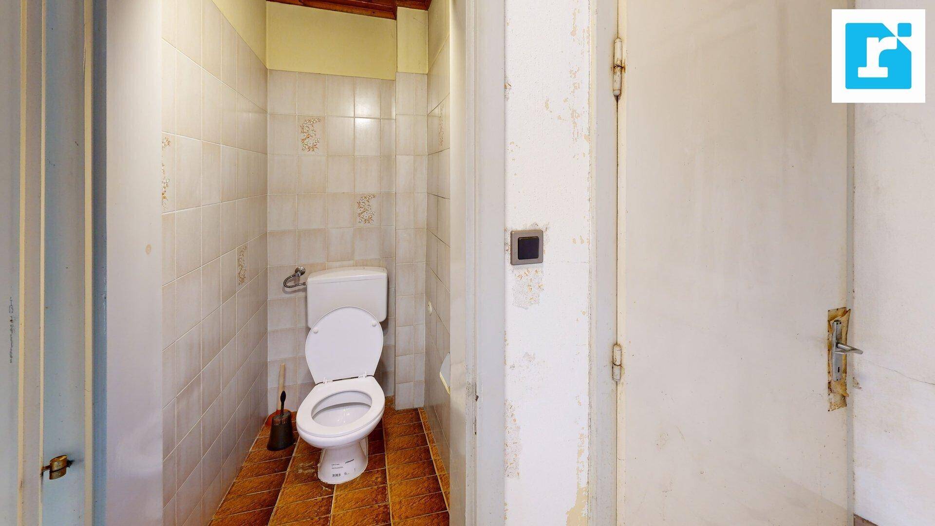 Einfache Toilette mit Fliesen und Holzdecke, in renovierungsbedürftigem Zustand.