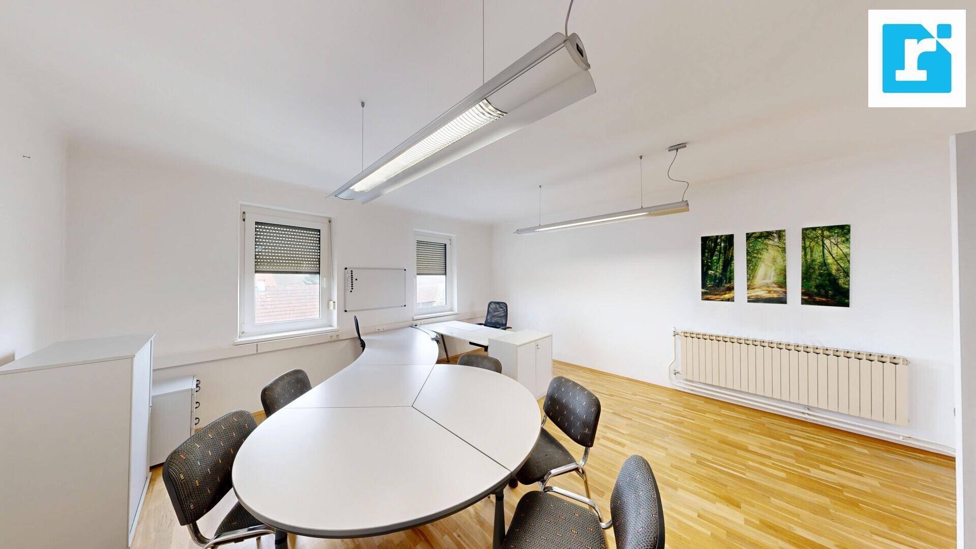 Geräumiges Büro mit Holzboden, einem großen Besprechungstisch und mehreren Fenstern mit Rollläden.