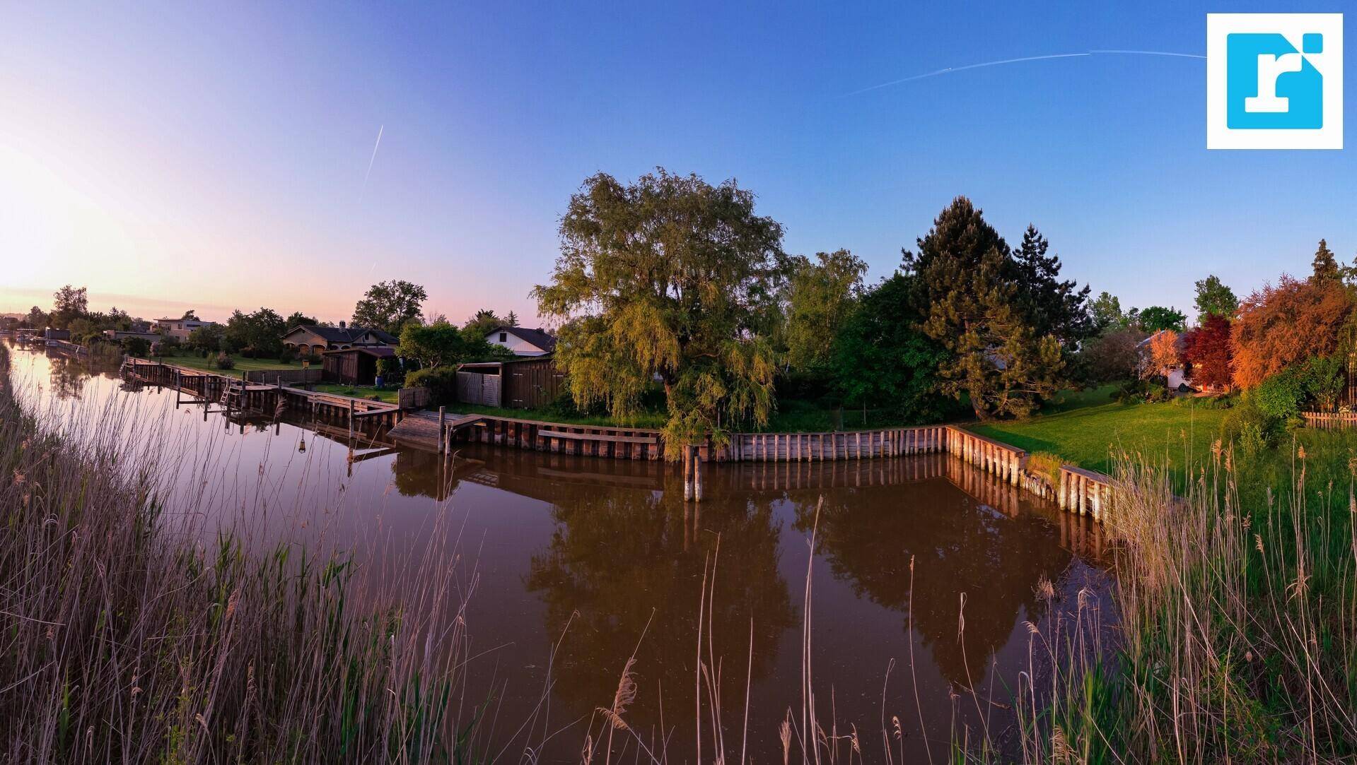 Malerischer Kanal mit Häusern am Ufer und üppiger Vegetation unter einem weiten Himmel.