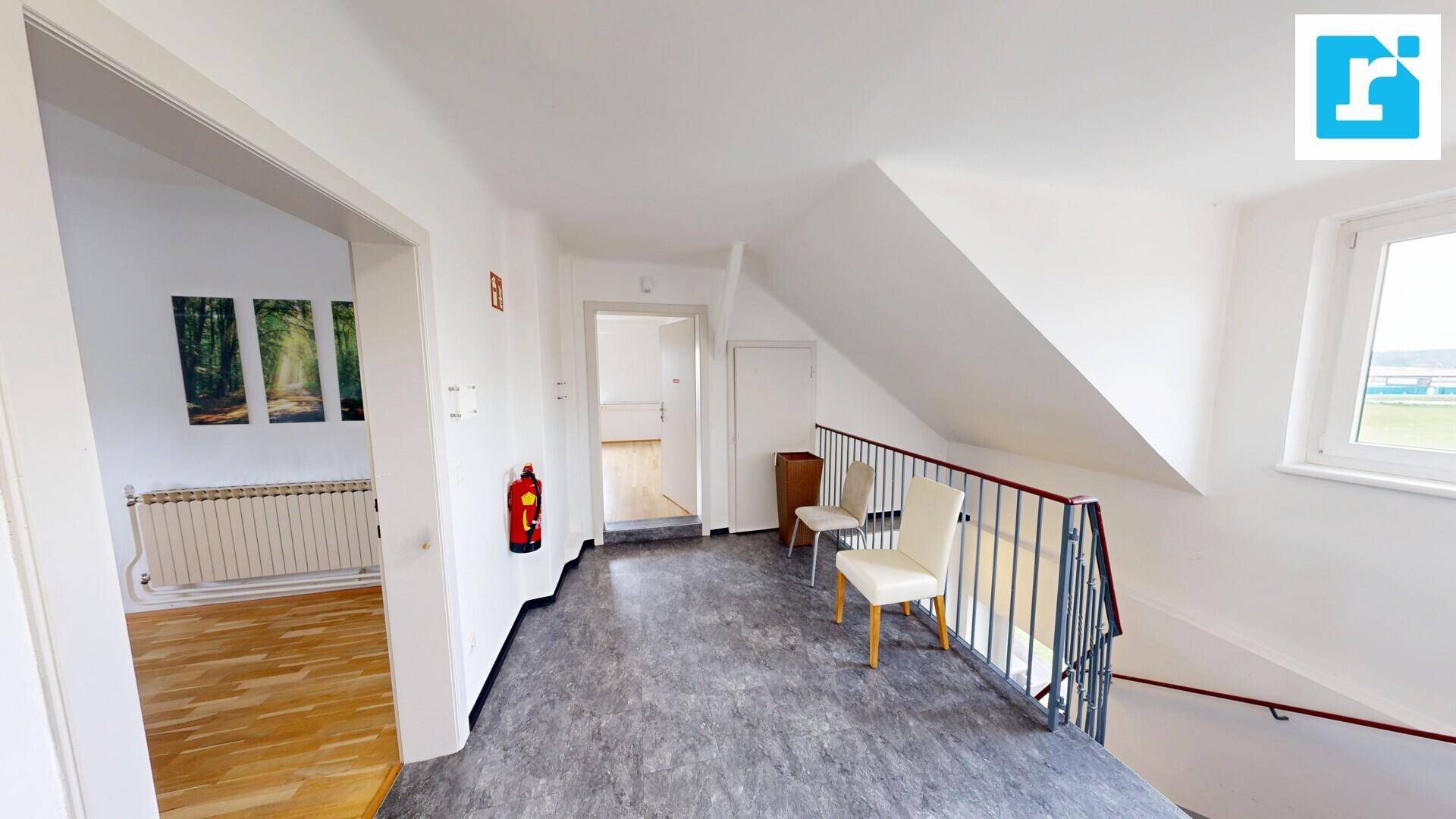Flur im Obergeschoss mit grauem Boden, einem Geländer und Zugang zu weiteren Räumen unter einem leicht schrägen Dach.
