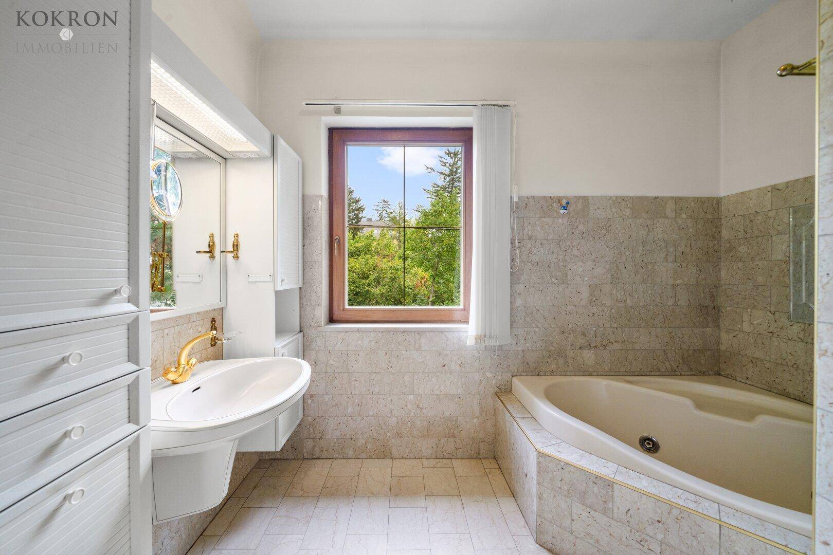 Badezimmer mit Eckbadewanne, Waschbecken und Fenster mit Blick ins Grüne.