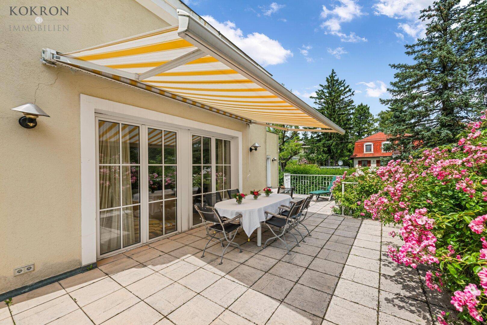 Große geflieste Terrasse mit Esstisch, Markise und blühenden Rosensträuchern im Garten.