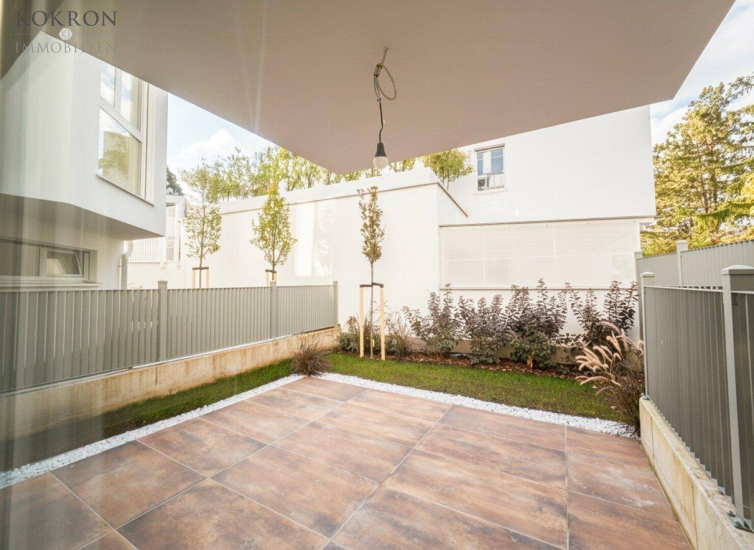 Kleine, gepflasterte Terrasse mit Kiesrand und junger Bepflanzung, umgeben von einer modernen Zaunanlage.